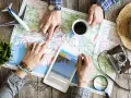 Pianificare viaggi senza agenzia: mani che studiano una mappa, un tablet con sito di viaggi, aereo giocattolo e macchina fotografica vintage.