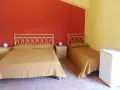 Camera semplice in un albergo una stella: due letti singoli e un letto matrimoniale con copriletti color ocra, pareti rosse e gialle, comodini e un piccolo frigorifero.