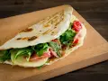 Piadina ricetta: focaccia ripiena di prosciutto, formaggio, rucola e pomodorini su tagliere di legno.