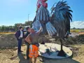 Operai al lavoro su un imponente gallo romagnolo in metallo, un'opera d'arte che domina il paesaggio.