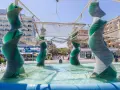 Sculture a spirale in vetro verde in una fontana a Riccione, con edifici e persone sullo sfondo.