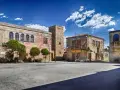 Piazza con palazzi fascisti, un torrione in pietra e alberi verdi sotto un cielo azzurro con nuvole bianche.