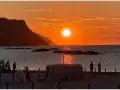 Un magico tramonto a Pesaro, con il sole che infuoca il mare e le colline sullo sfondo. Persone godono della spiaggia.