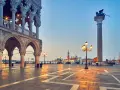L'alba illumina il colonnato del Palazzo Ducale, proiettando lunghe ombre sulla piazza. In lontananza, il leone alato veglia su Piazza San Marco.