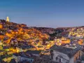 Un viaggio on the road in Basilicata, con Matera illuminata al crepuscolo. Case antiche e chiese rupestri brillano di luci calde.