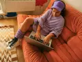 Ragazza con cuffie e cappellino viola lavora al laptop su un divano arancione. Si può andare in hotel a 16 anni, ma lei preferisce casa.