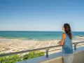 Vista panoramica di una spiaggia affollata a Cattolica, con ombrelloni e lettini disposti ordinatamente. Una donna ammira il mare azzurro da un balcone.