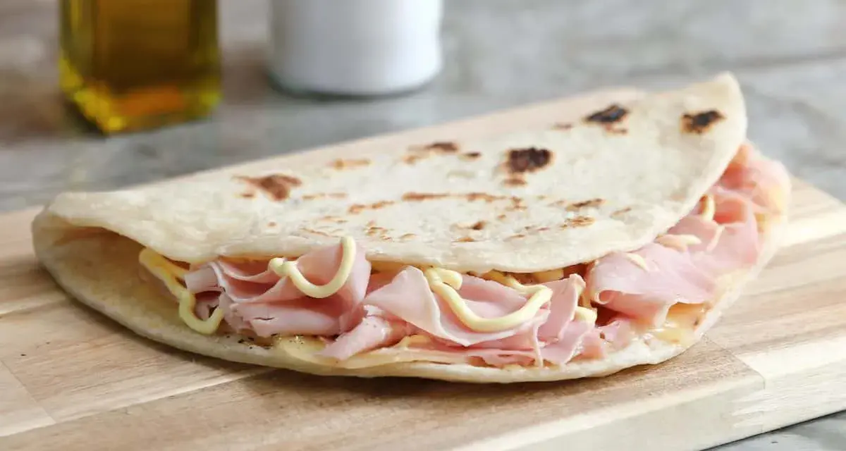 Piadina sfogliata romagnola ripiena di prosciutto cotto e formaggio, su tagliere di legno.