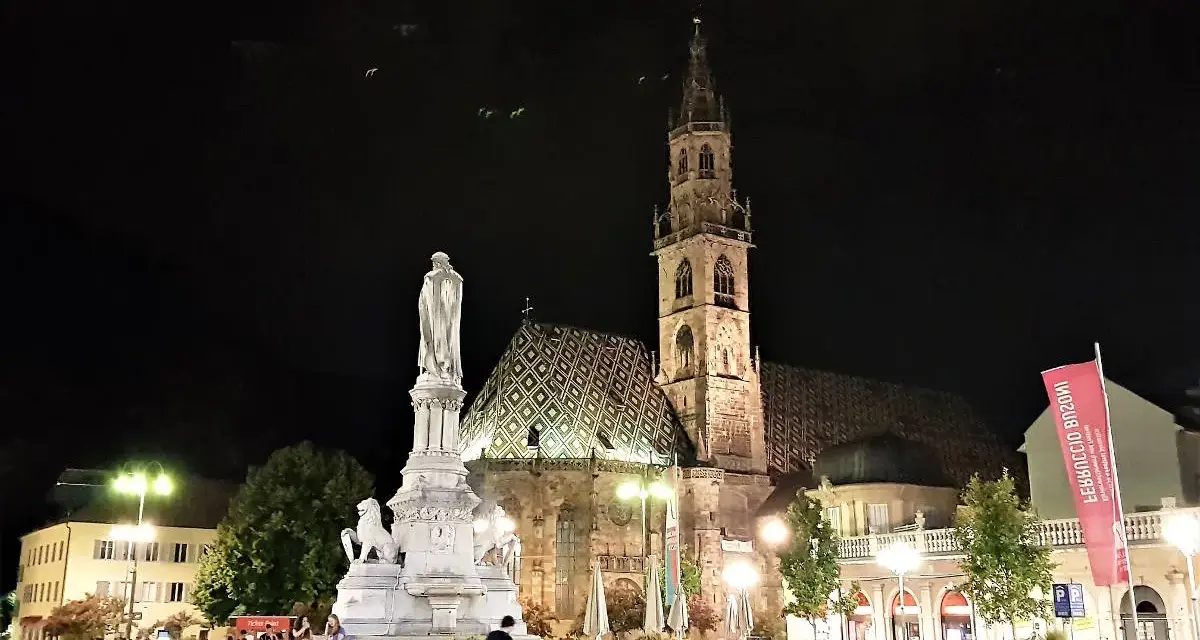 Piazza Walther di Bolzano di notte: la cattedrale, il monumento e la bandiera rossa. Un'idea di cosa vedere a Bolzano in un giorno.
