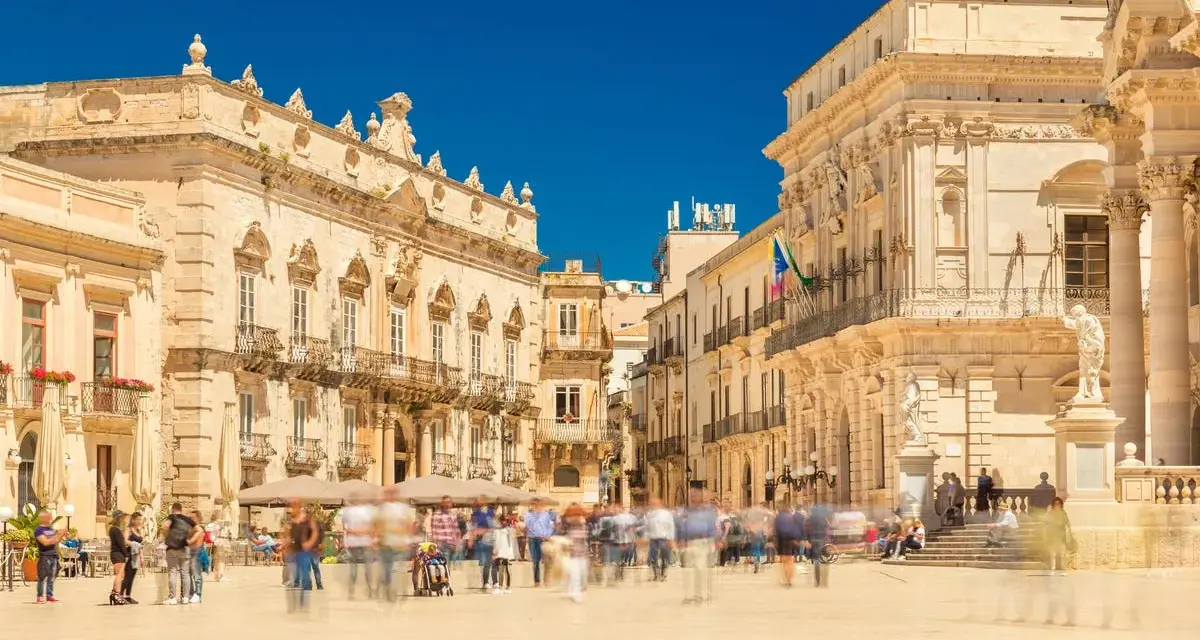 Piazza affollata con edifici storici. La gente si muove rapidamente, quasi sfocata, mentre si valuta la **classificazione stelle hotel** per un soggiorno in questa splendida cornice.