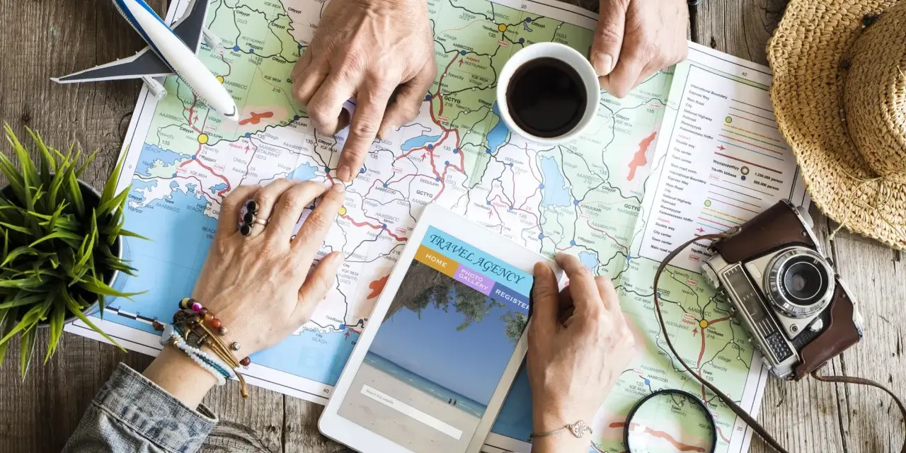 Pianificare viaggi senza agenzia: mani che studiano una mappa, un tablet con sito di viaggi, aereo giocattolo e macchina fotografica vintage.