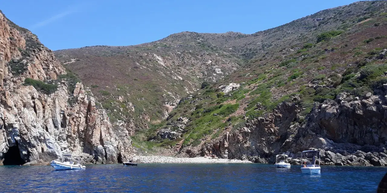 Barche a vela nella cala del corvo, con scogliere rocciose e vegetazione mediterranea.