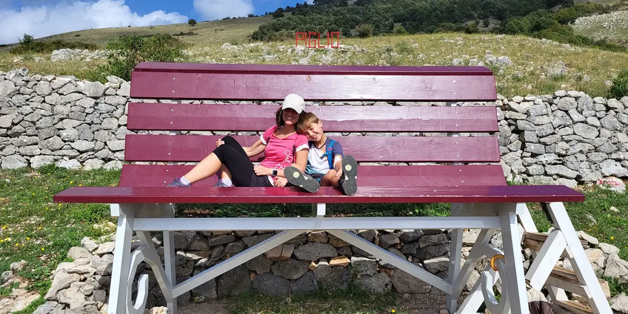 Gita domenicale con bambini nel Lazio: mamma e figlio si godono il panorama su una panchina gigante, con montagne e cielo azzurro sullo sfondo.