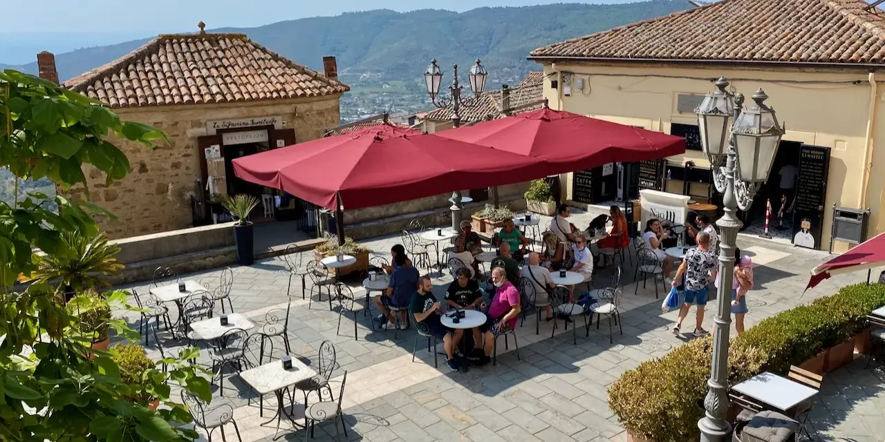 Piazza soleggiata a Castellabate con tavolini all'aperto e gente che si gode il momento. La scritta "qui non si muore" invita a rilassarsi.