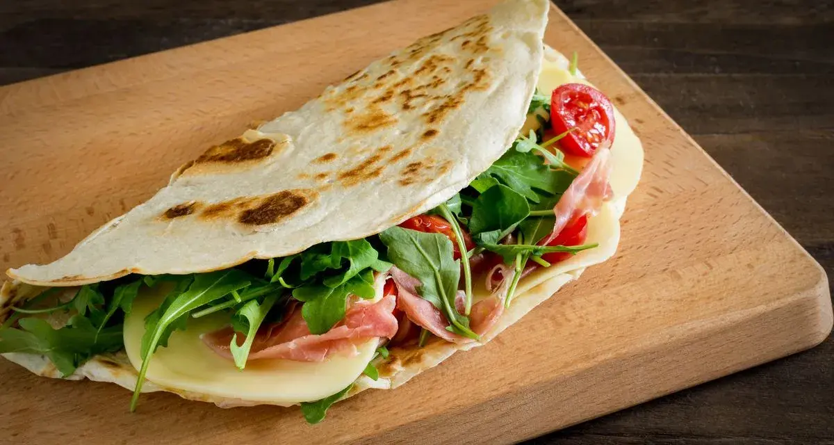 Piadina ricetta: focaccia ripiena di prosciutto, formaggio, rucola e pomodorini su tagliere di legno.