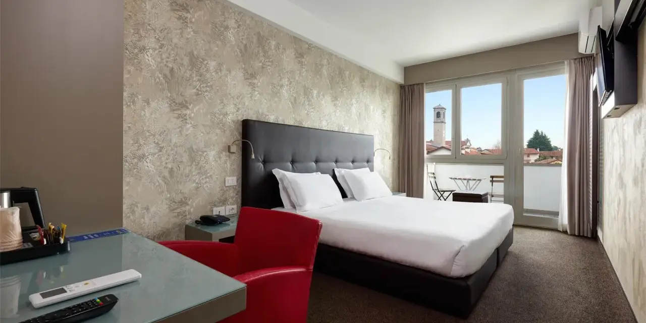Camera moderna con letto matrimoniale, scrivania in vetro, poltrona rossa e balcone con vista. I prezzi su Booking sono a persona.