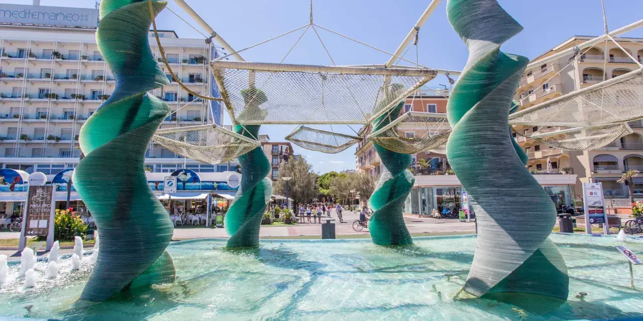 Sculture a spirale in vetro verde in una fontana a Riccione, con edifici e persone sullo sfondo.