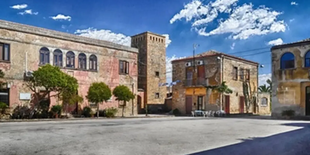 Piazza con palazzi fascisti, un torrione in pietra e alberi verdi sotto un cielo azzurro con nuvole bianche.