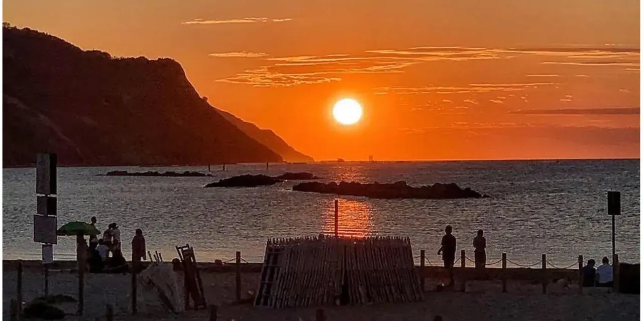 Un magico tramonto a Pesaro, con il sole che infuoca il mare e le colline sullo sfondo. Persone godono della spiaggia.