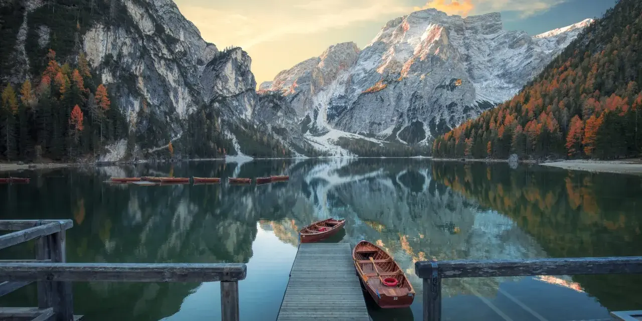 Lago di Braies, con montagne innevate e boschi autunnali riflessi sull'acqua. Per sapere come arrivare al lago di Braies in treno da Roma, cerca online.