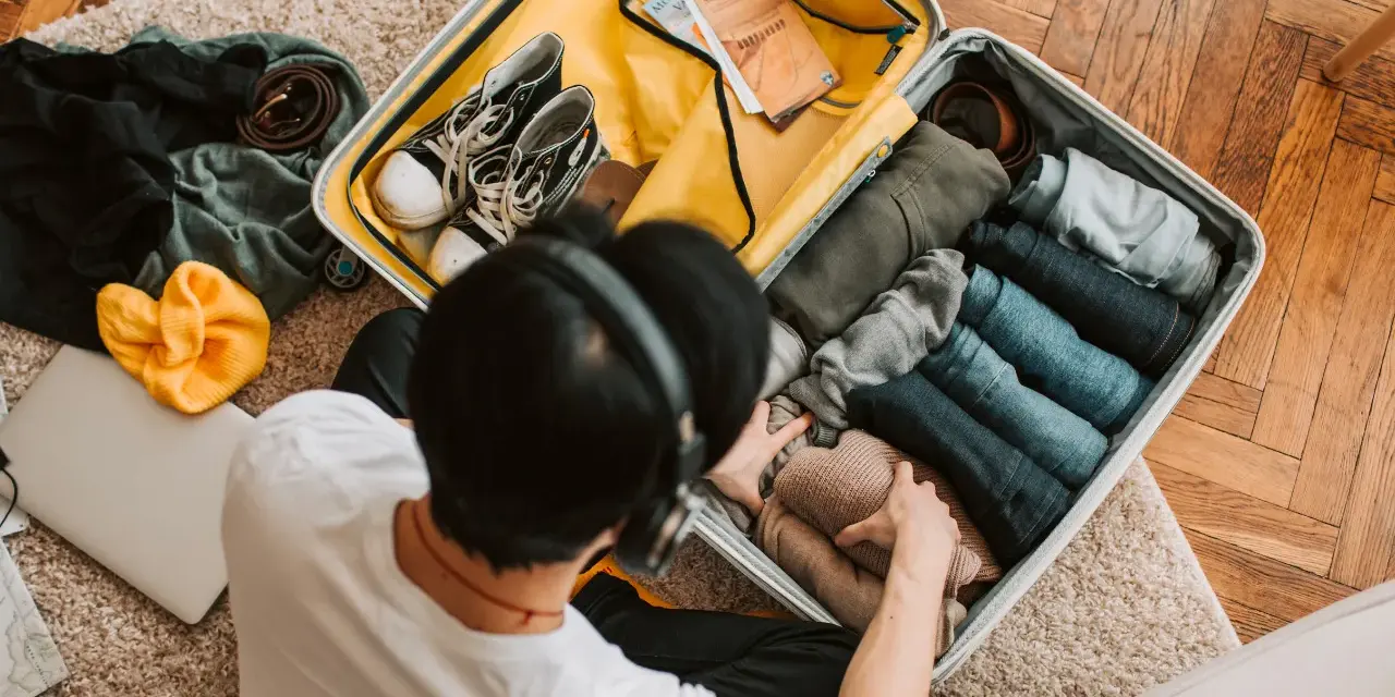 Preparazione per la gita: valigia aperta con vestiti piegati, scarpe, cintura, cappello giallo e laptop.