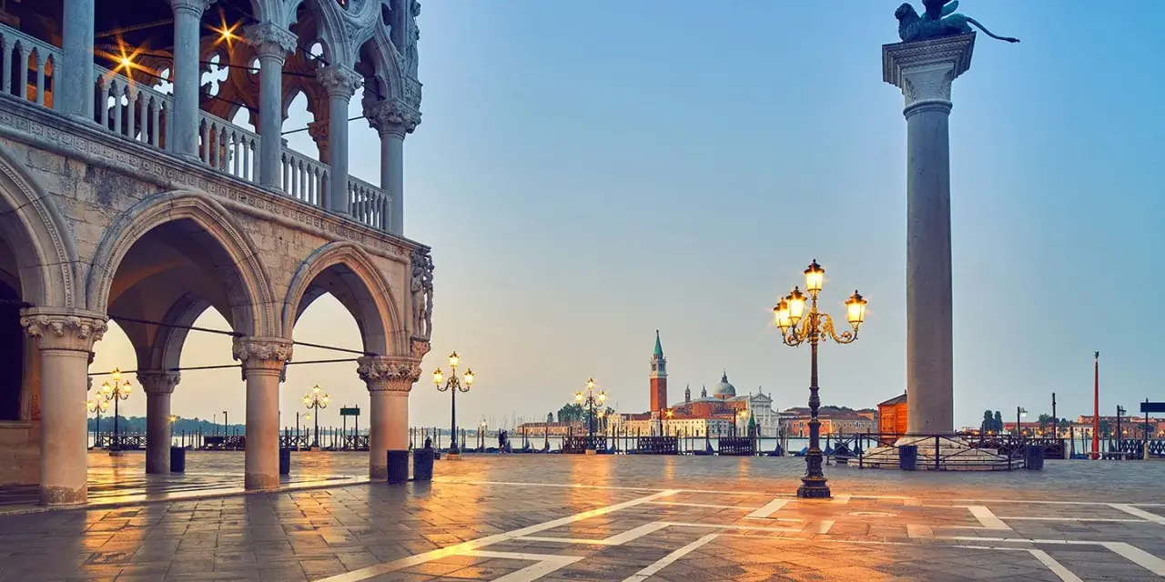 L'alba illumina il colonnato del Palazzo Ducale, proiettando lunghe ombre sulla piazza. In lontananza, il leone alato veglia su Piazza San Marco.