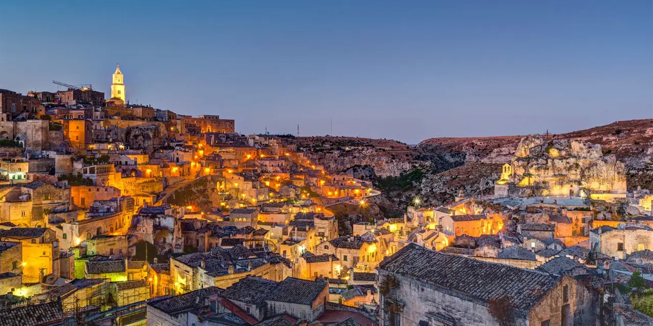 Un viaggio on the road in Basilicata, con Matera illuminata al crepuscolo. Case antiche e chiese rupestri brillano di luci calde.