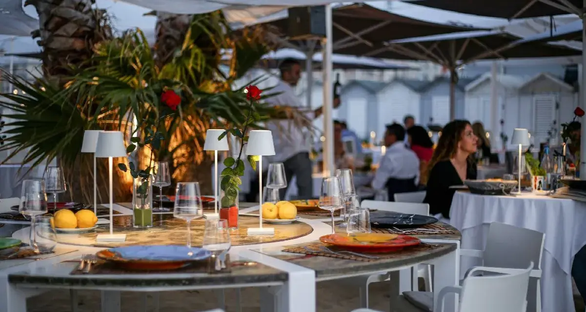 Tavoli apparecchiati sulla spiaggia di Riccione, con limoni, rose rosse e piccole lampade. Atmosfera serale, perfetta per una cena di manna.
