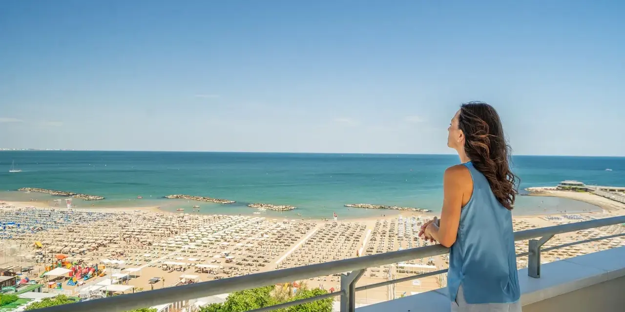 Vista panoramica di una spiaggia affollata a Cattolica, con ombrelloni e lettini disposti ordinatamente. Una donna ammira il mare azzurro da un balcone.
