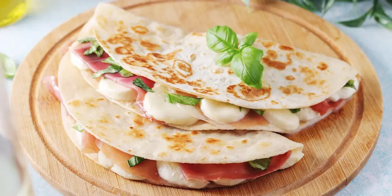 Piadina romagnola ripiena di prosciutto crudo, mozzarella e basilico, servita su un tagliere di legno.
