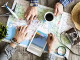 Pianificare viaggi senza agenzia: mani che studiano una mappa, un tablet con sito di viaggi, aereo giocattolo e macchina fotografica vintage.
