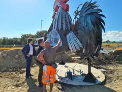 Operai al lavoro su un imponente gallo romagnolo in metallo, un'opera d'arte che domina il paesaggio.
