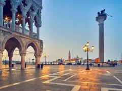 L'alba illumina il colonnato del Palazzo Ducale, proiettando lunghe ombre sulla piazza. In lontananza, il leone alato veglia su Piazza San Marco.