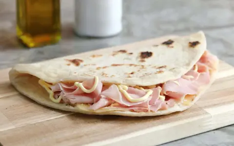 Piadina sfogliata romagnola ripiena di prosciutto cotto e formaggio, su tagliere di legno.