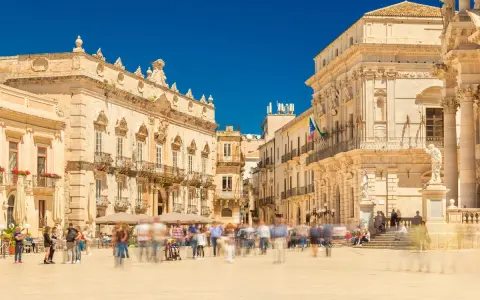 Piazza affollata con edifici storici. La gente si muove rapidamente, quasi sfocata, mentre si valuta la **classificazione stelle hotel** per un soggiorno in questa splendida cornice.