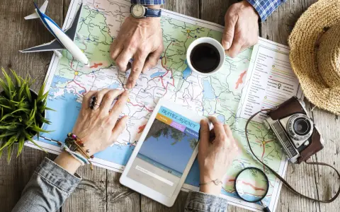 Pianificare viaggi senza agenzia: mani che studiano una mappa, un tablet con sito di viaggi, aereo giocattolo e macchina fotografica vintage.