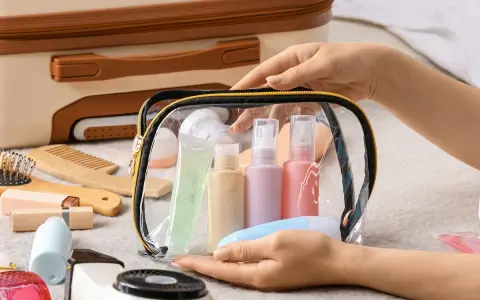 Mani che preparano cosa mettere nel beauty case: flaconi, gel e accessori da viaggio in una trousse trasparente.