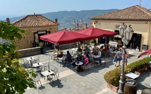Piazza soleggiata a Castellabate con tavolini all'aperto e gente che si gode il momento. La scritta "qui non si muore" invita a rilassarsi.