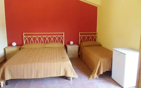 Camera semplice in un albergo una stella: due letti singoli e un letto matrimoniale con copriletti color ocra, pareti rosse e gialle, comodini e un piccolo frigorifero.