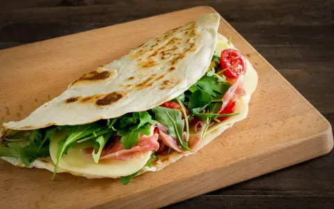 Piadina ricetta: focaccia ripiena di prosciutto, formaggio, rucola e pomodorini su tagliere di legno.