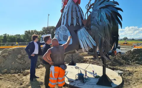 Operai al lavoro su un imponente gallo romagnolo in metallo, un'opera d'arte che domina il paesaggio.