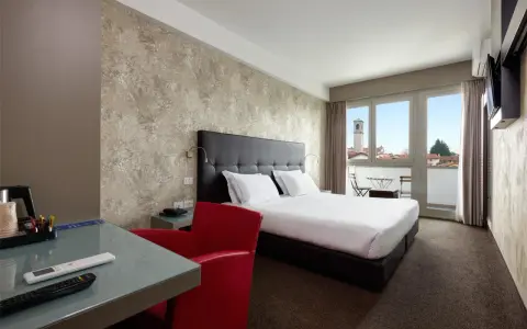 Camera moderna con letto matrimoniale, scrivania in vetro, poltrona rossa e balcone con vista. I prezzi su Booking sono a persona.
