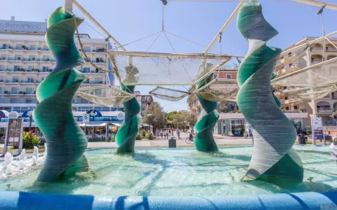 Sculture a spirale in vetro verde in una fontana a Riccione, con edifici e persone sullo sfondo.
