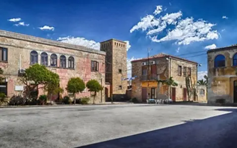 Piazza con palazzi fascisti, un torrione in pietra e alberi verdi sotto un cielo azzurro con nuvole bianche.