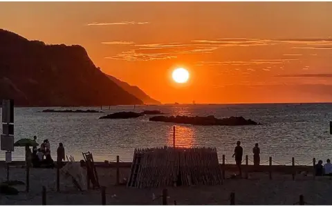 Un magico tramonto a Pesaro, con il sole che infuoca il mare e le colline sullo sfondo. Persone godono della spiaggia.