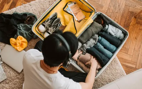 Preparazione per la gita: valigia aperta con vestiti piegati, scarpe, cintura, cappello giallo e laptop.