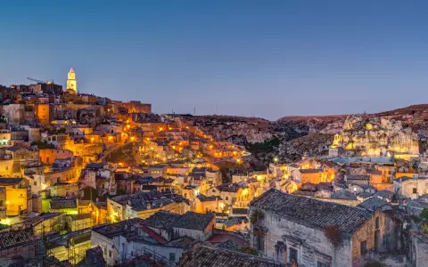 Un viaggio on the road in Basilicata, con Matera illuminata al crepuscolo. Case antiche e chiese rupestri brillano di luci calde.