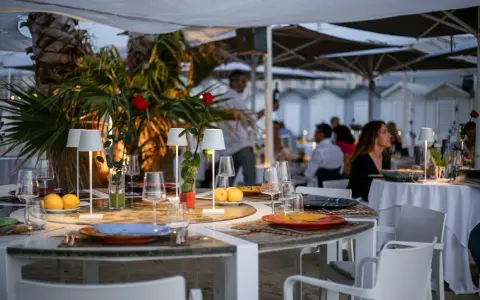 Tavoli apparecchiati sulla spiaggia di Riccione, con limoni, rose rosse e piccole lampade. Atmosfera serale, perfetta per una cena di manna.