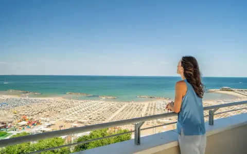 Vista panoramica di una spiaggia affollata a Cattolica, con ombrelloni e lettini disposti ordinatamente. Una donna ammira il mare azzurro da un balcone.