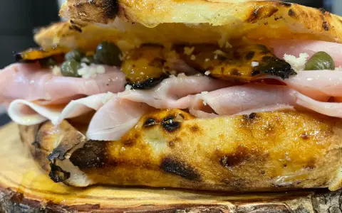 Un panino gourmet con mortadella, capperi e pomodori secchi, un vero re artù a Lecce, servito su un tagliere di legno.